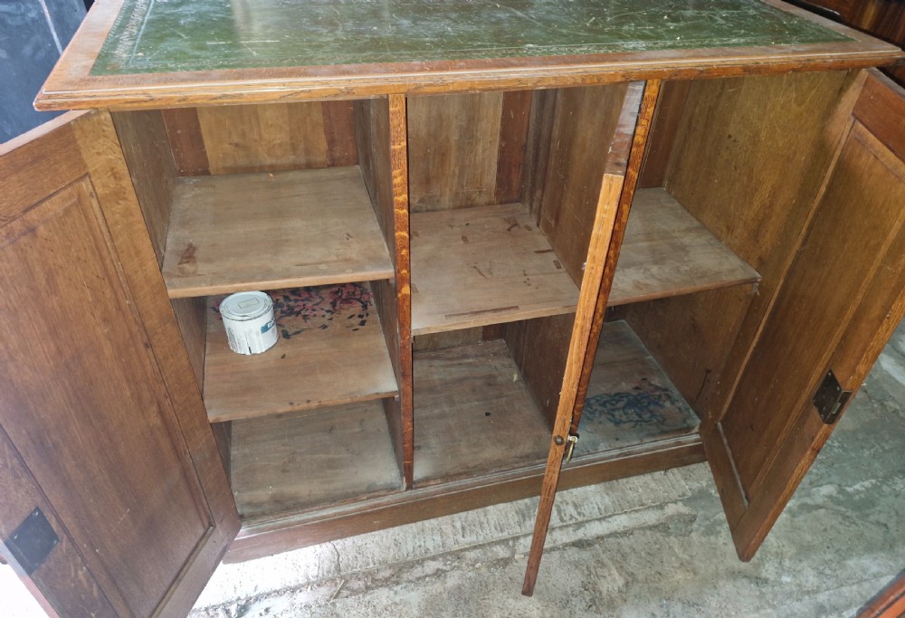 An antique golden oak cupboard featuring three panelled doors, brass ring handles, and an inset green leather top surface. View II