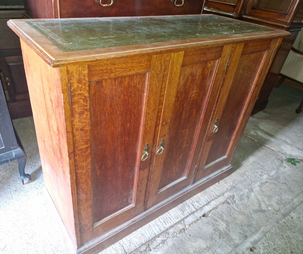 An antique golden oak cupboard featuring three panelled doors, brass ring handles, and an inset green leather top surface. View IV