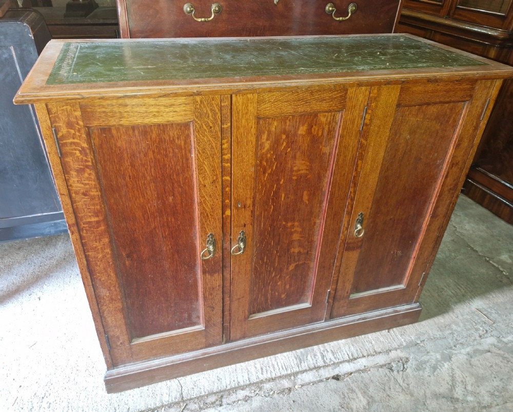 An antique golden oak cupboard featuring three panelled doors, brass ring handles, and an inset green leather top surface. View V