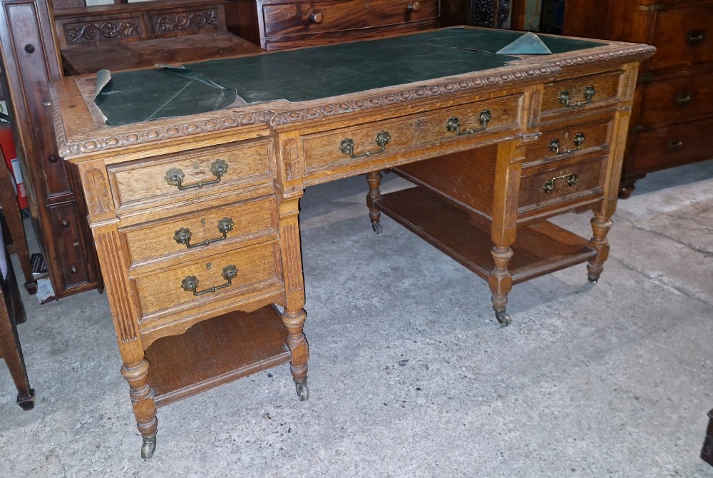 An Edwardian carved oak kneehole pedestal desk featuring a green leather writing insert, multiple drawers with brass swing handles, carved borders, and turned legs set on castors. View II