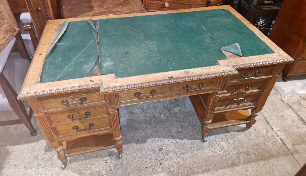 An Edwardian carved oak kneehole pedestal desk featuring a green leather writing insert, multiple drawers with brass swing handles, carved borders, and turned legs set on castors. View V