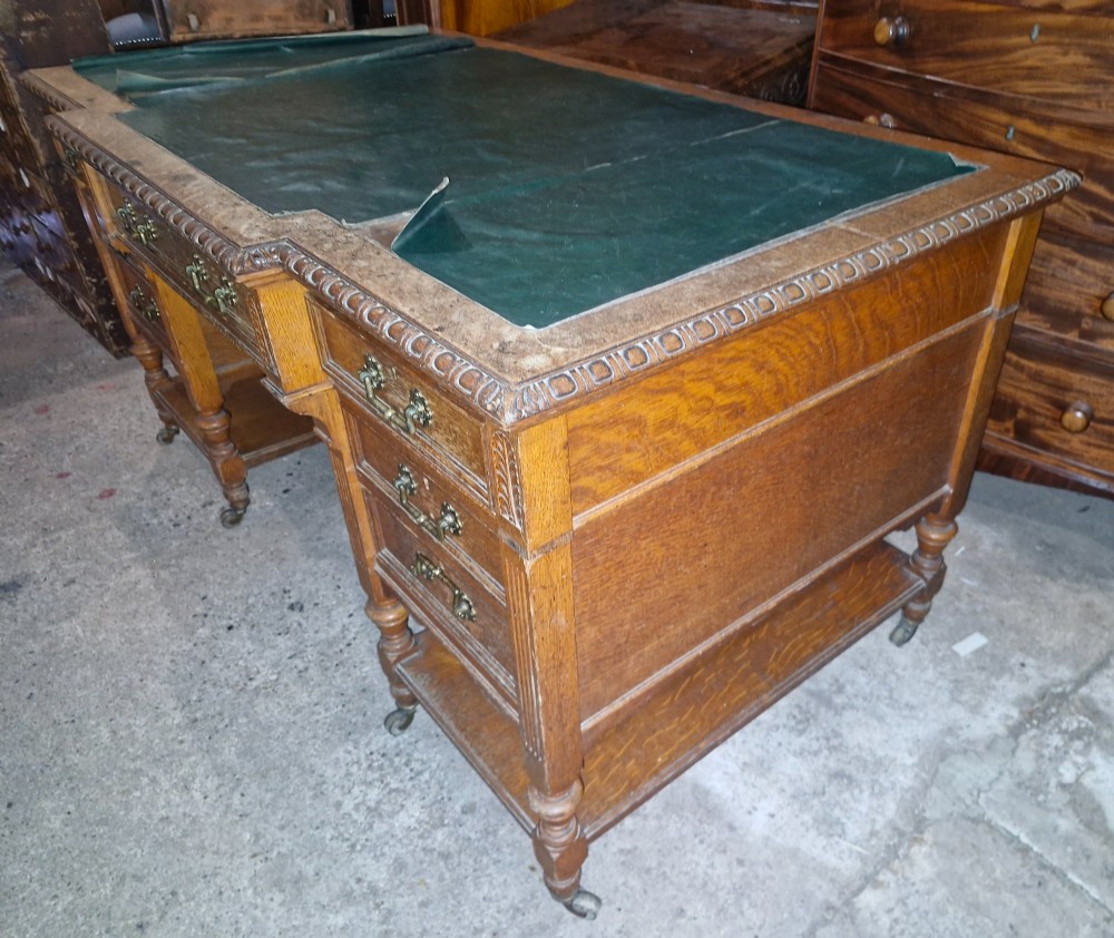 An Edwardian carved oak kneehole pedestal desk featuring a green leather writing insert, multiple drawers with brass swing handles, carved borders, and turned legs set on castors. View VII