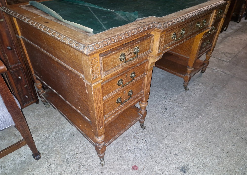 An Edwardian carved oak kneehole pedestal desk featuring a green leather writing insert, multiple drawers with brass swing handles, carved borders, and turned legs set on castors. View VIII