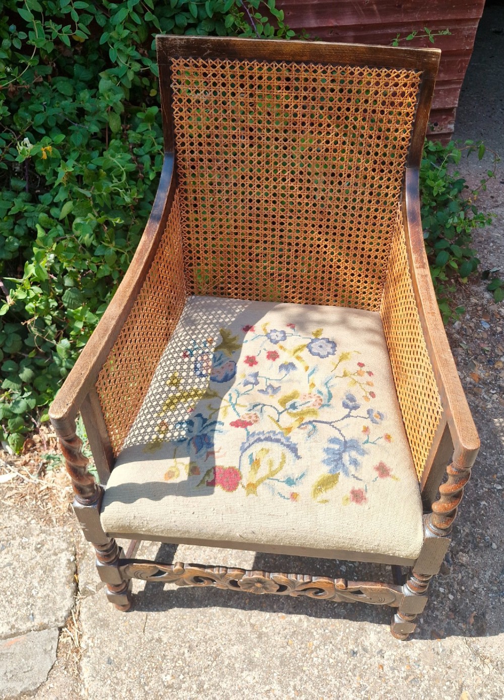 A dark oak bergere armchair featuring woven cane back and side panels, barley twist arm supports, a carved front stretcher, and a floral tapestry seat cushion. View III