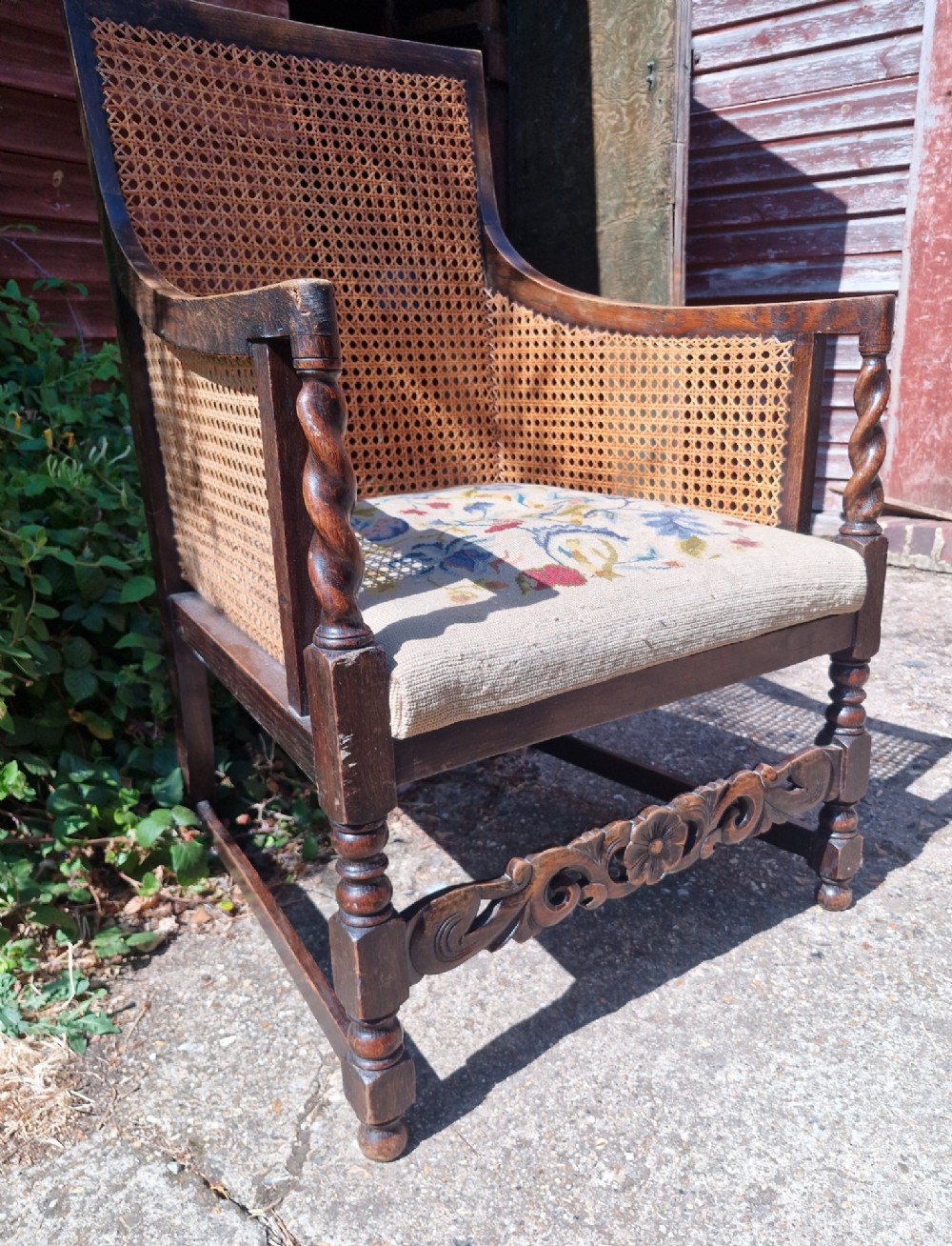 A dark oak bergere armchair featuring woven cane back and side panels, barley twist arm supports, a carved front stretcher, and a floral tapestry seat cushion. View IV