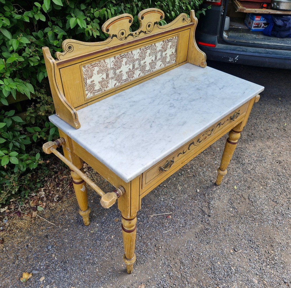 A late Victorian yellow painted washstand featuring a white marble top, ceramic tiled splashback with floral motifs, a single drawer, and turned legs. View V
