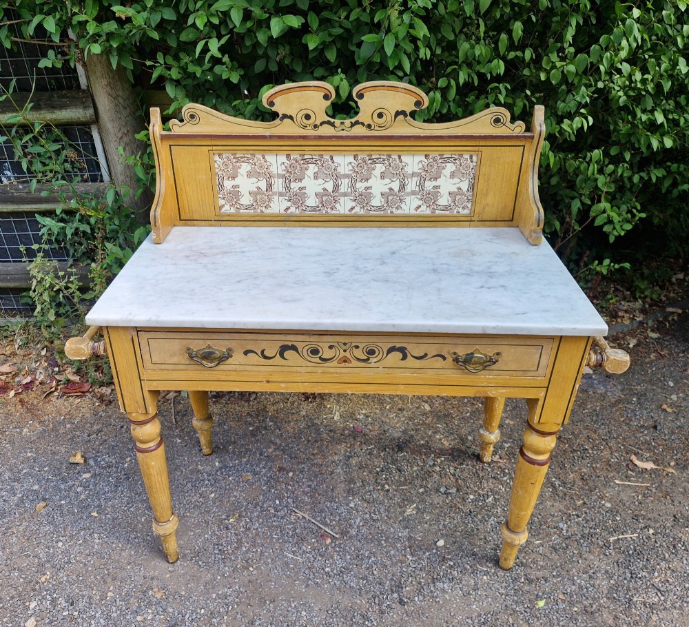 A late Victorian yellow painted washstand featuring a white marble top, ceramic tiled splashback with floral motifs, a single drawer, and turned legs. View VI