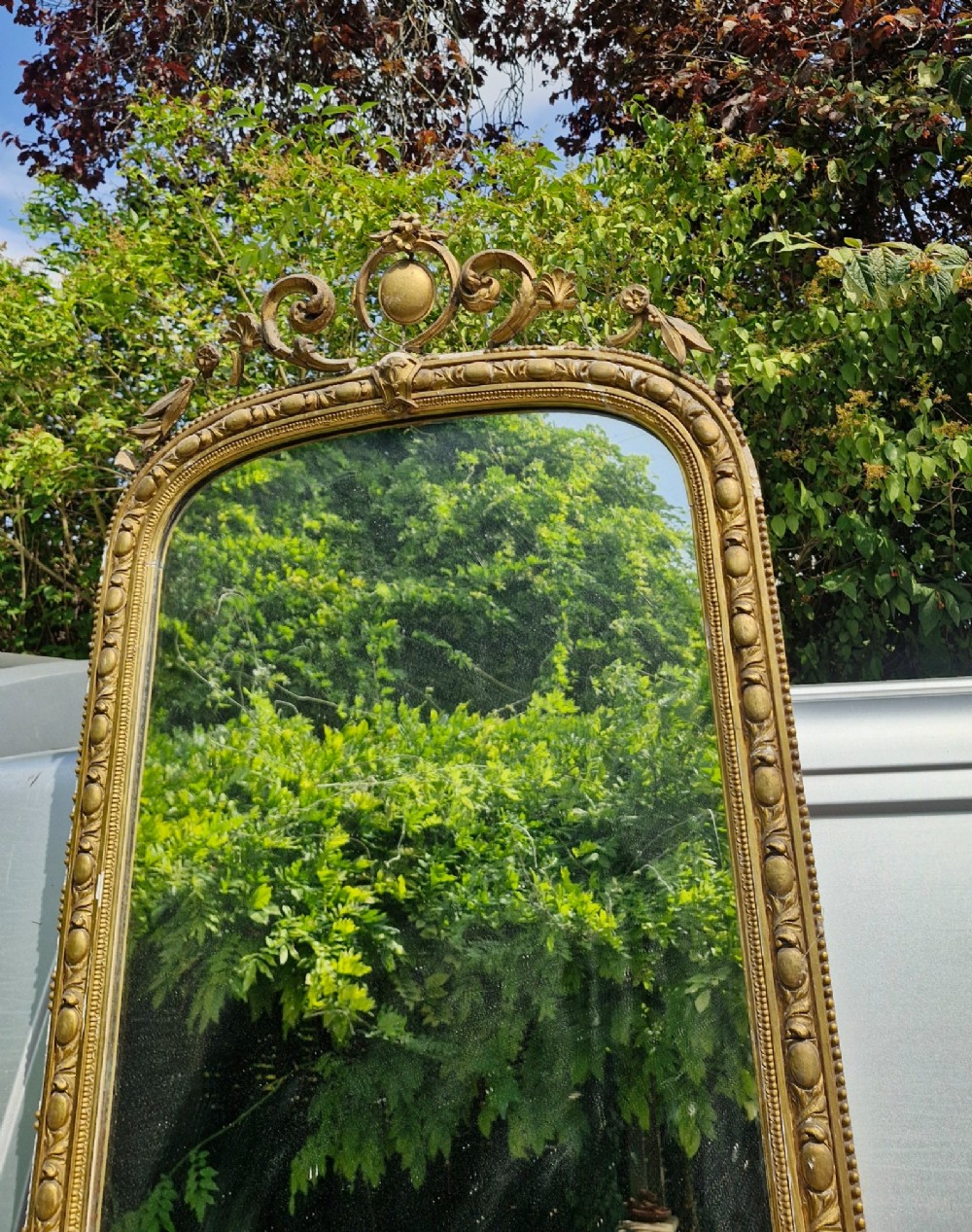 A tall Victorian mirror featuring an arched giltwood frame decorated with bead moulding and an ornate cresting motif at the top. View V