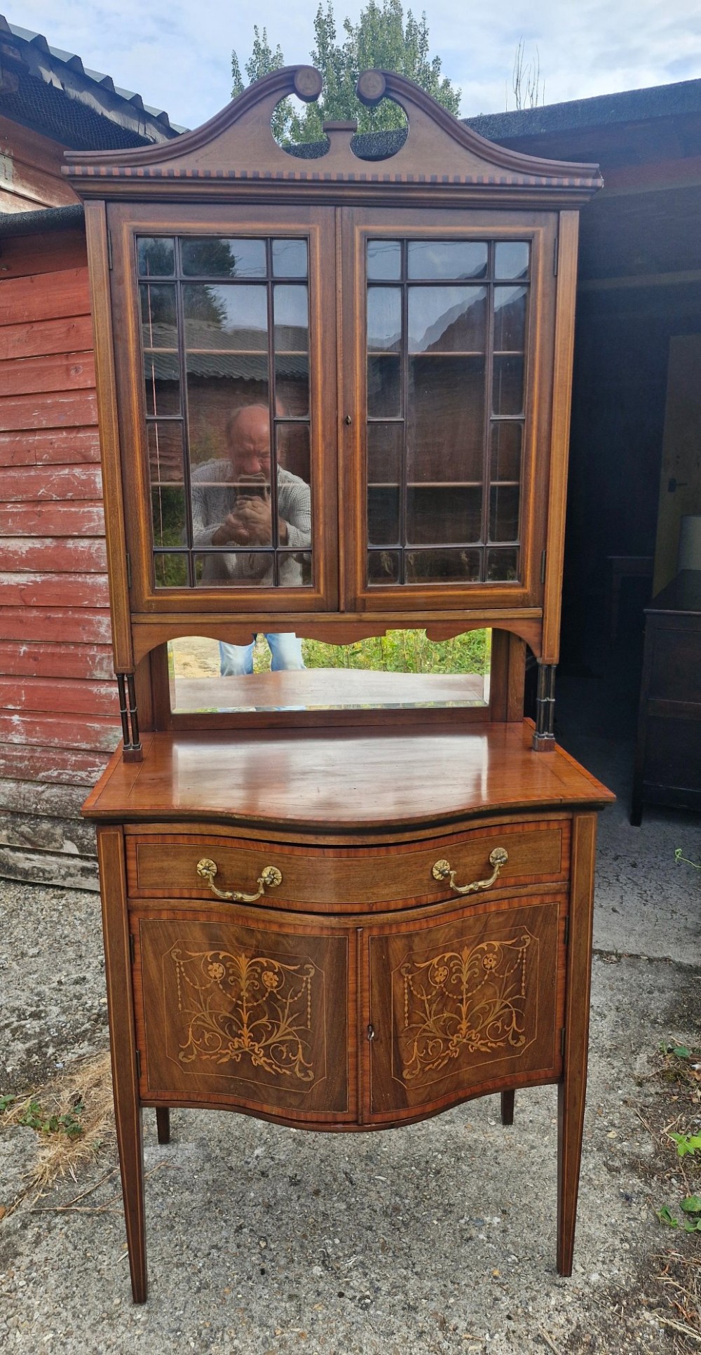 An Edwardian inlaid mahogany two-part display cabinet featuring a swan-neck pediment, glazed upper doors, a mirrored splashback, and lower cupboard doors with floral marquetry.