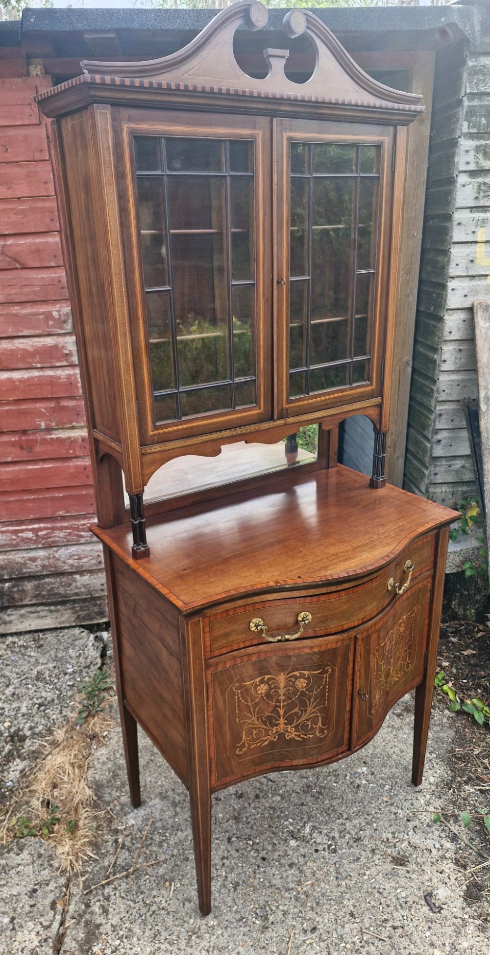 An Edwardian inlaid mahogany two-part display cabinet featuring a swan-neck pediment, glazed upper doors, a mirrored splashback, and lower cupboard doors with floral marquetry. View III