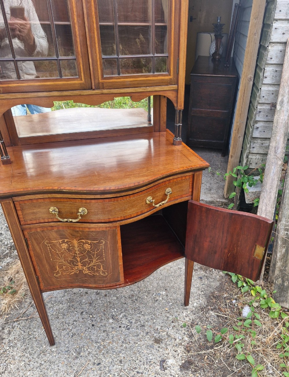 An Edwardian inlaid mahogany two-part display cabinet featuring a swan-neck pediment, glazed upper doors, a mirrored splashback, and lower cupboard doors with floral marquetry. View IV