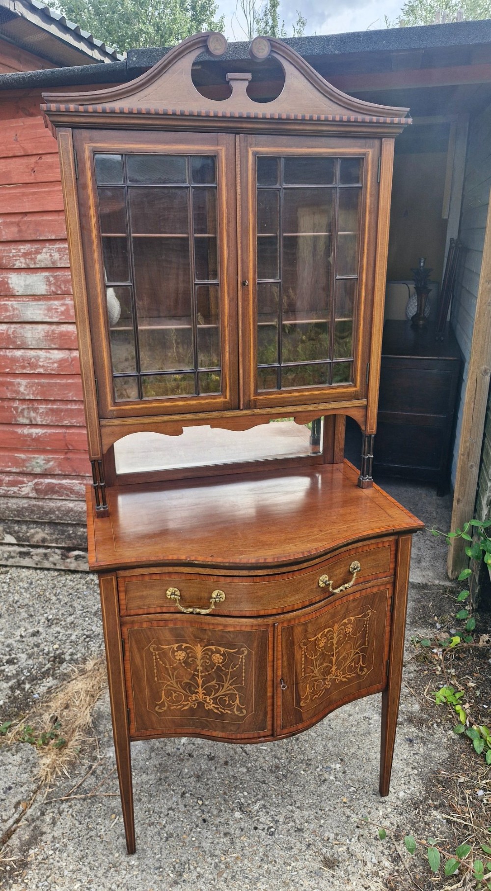 An Edwardian inlaid mahogany two-part display cabinet featuring a swan-neck pediment, glazed upper doors, a mirrored splashback, and lower cupboard doors with floral marquetry. View XI