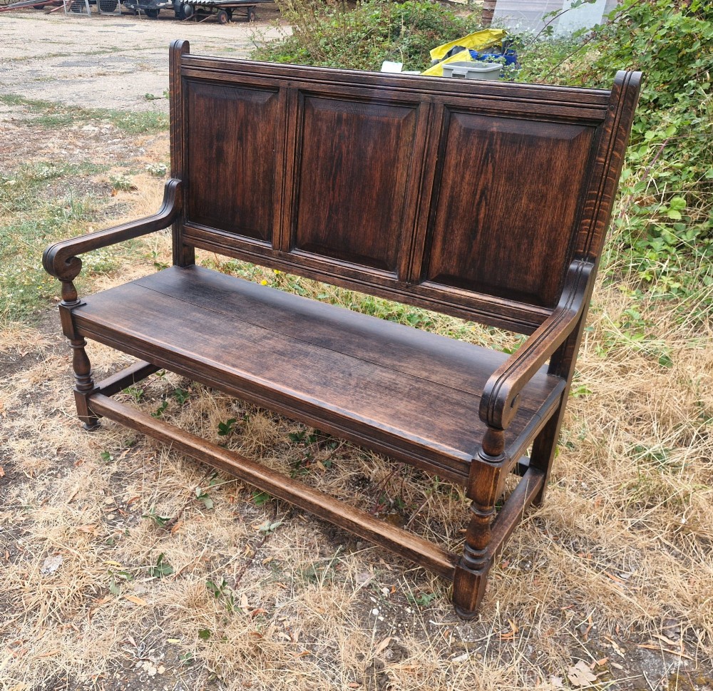 A dark oak hall bench featuring a triple-panelled backrest, scrolled armrests, turned front legs, and a solid wooden seat.