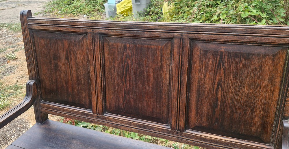 A dark oak hall bench featuring a triple-panelled backrest, scrolled armrests, turned front legs, and a solid wooden seat. View III