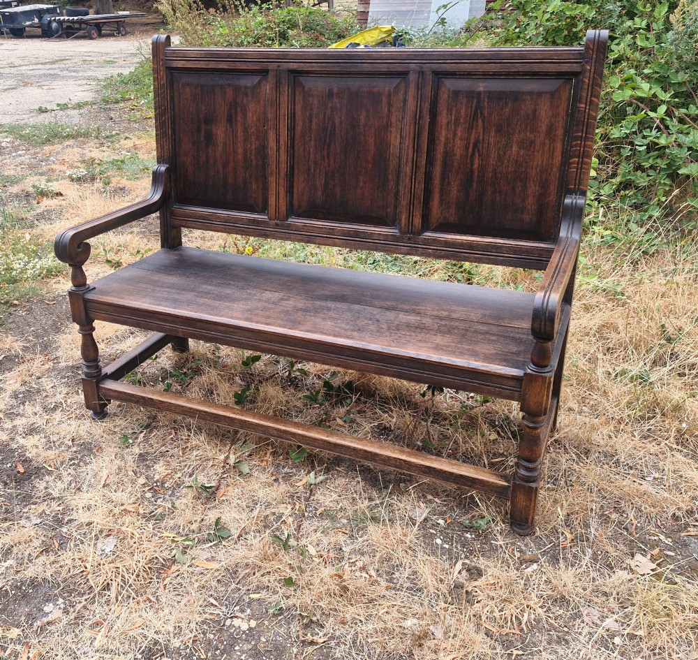 A dark oak hall bench featuring a triple-panelled backrest, scrolled armrests, turned front legs, and a solid wooden seat. View IV