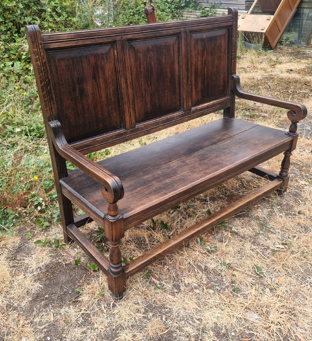 A dark oak hall bench featuring a triple-panelled backrest, scrolled armrests, turned front legs, and a solid wooden seat. View V