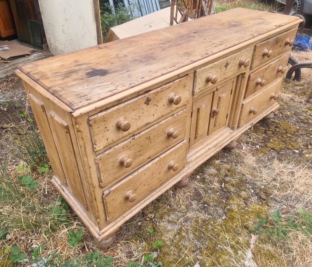 A wide Victorian pine dresser base featuring multiple drawers with turned wooden handles flanking a central double cupboard, resting on turned feet. View IV