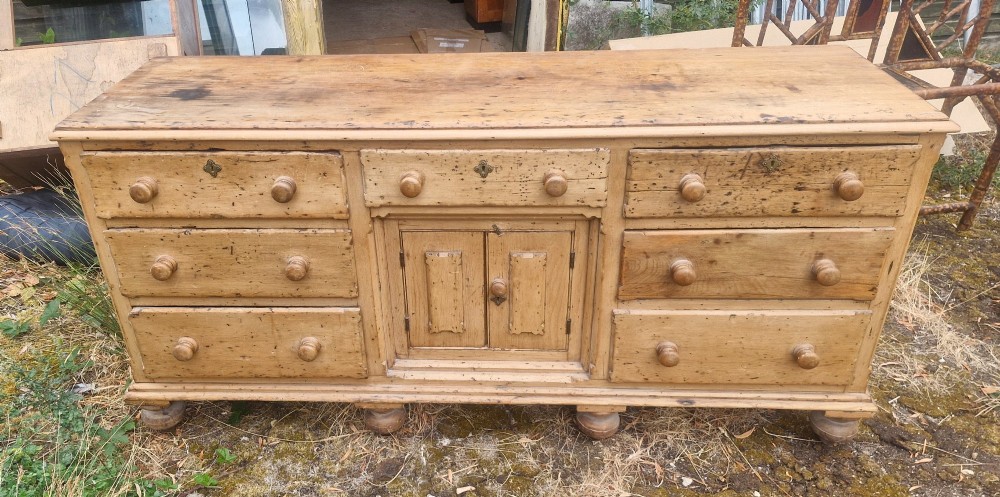 A wide Victorian pine dresser base featuring multiple drawers with turned wooden handles flanking a central double cupboard, resting on turned feet. View V