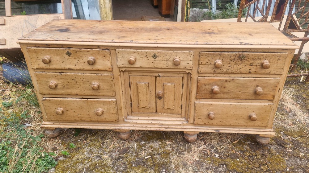 A wide Victorian pine dresser base featuring multiple drawers with turned wooden handles flanking a central double cupboard, resting on turned feet. View VI