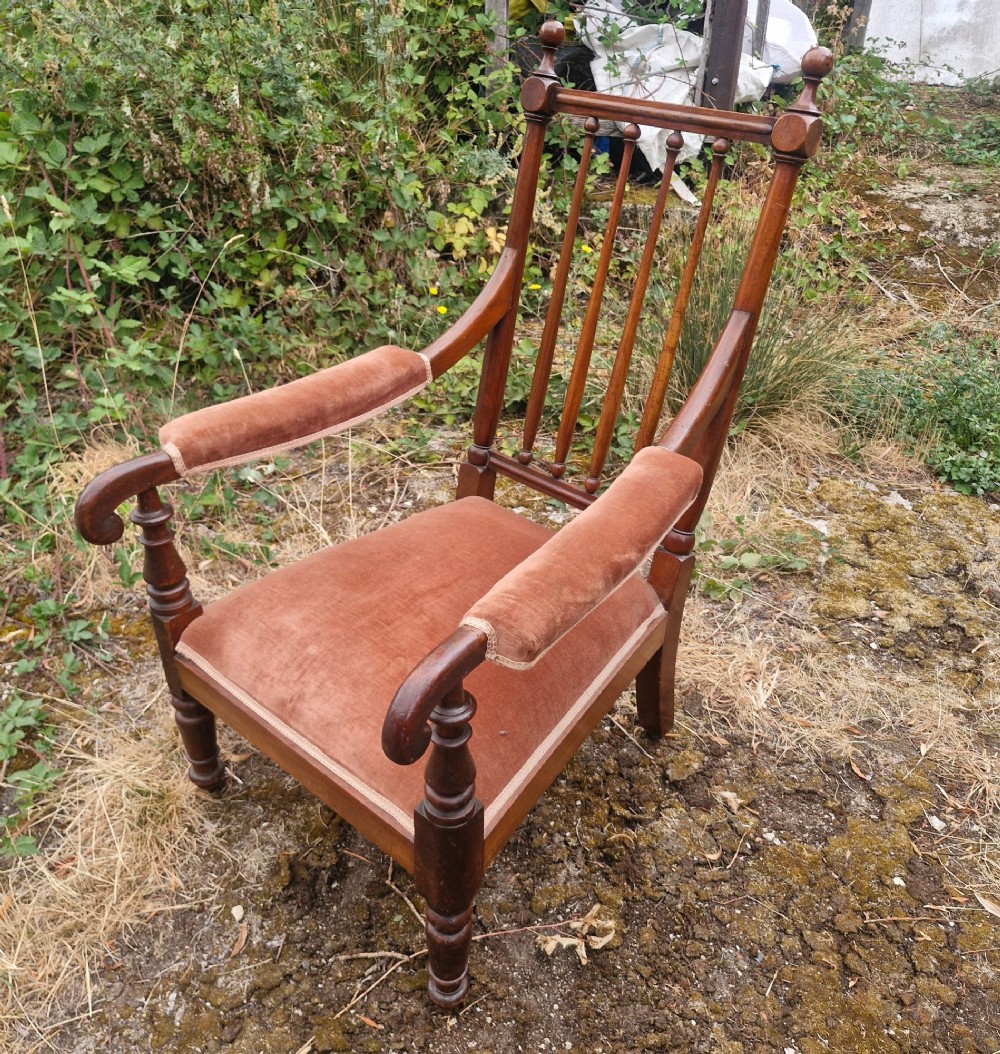 An early Victorian mahogany armchair featuring a spindle backrest, padded velvet armrests, a cushioned brown velvet seat, and turned wooden front legs.
