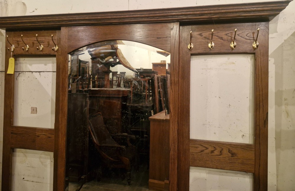 A large dark oak hall stand featuring a central arched mirror flanked by brass coat hooks, set above built-in storage boxes and a central trough base. View VI
