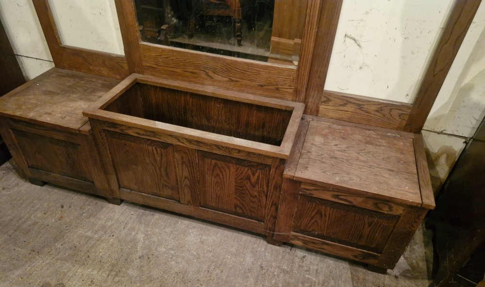 A large dark oak hall stand featuring a central arched mirror flanked by brass coat hooks, set above built-in storage boxes and a central trough base. View VII