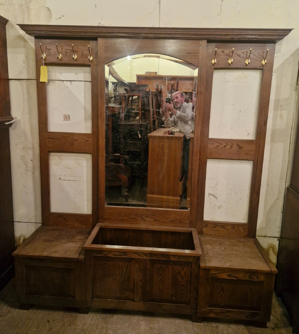 A large dark oak hall stand featuring a central arched mirror flanked by brass coat hooks, set above built-in storage boxes and a central trough base. View VIII