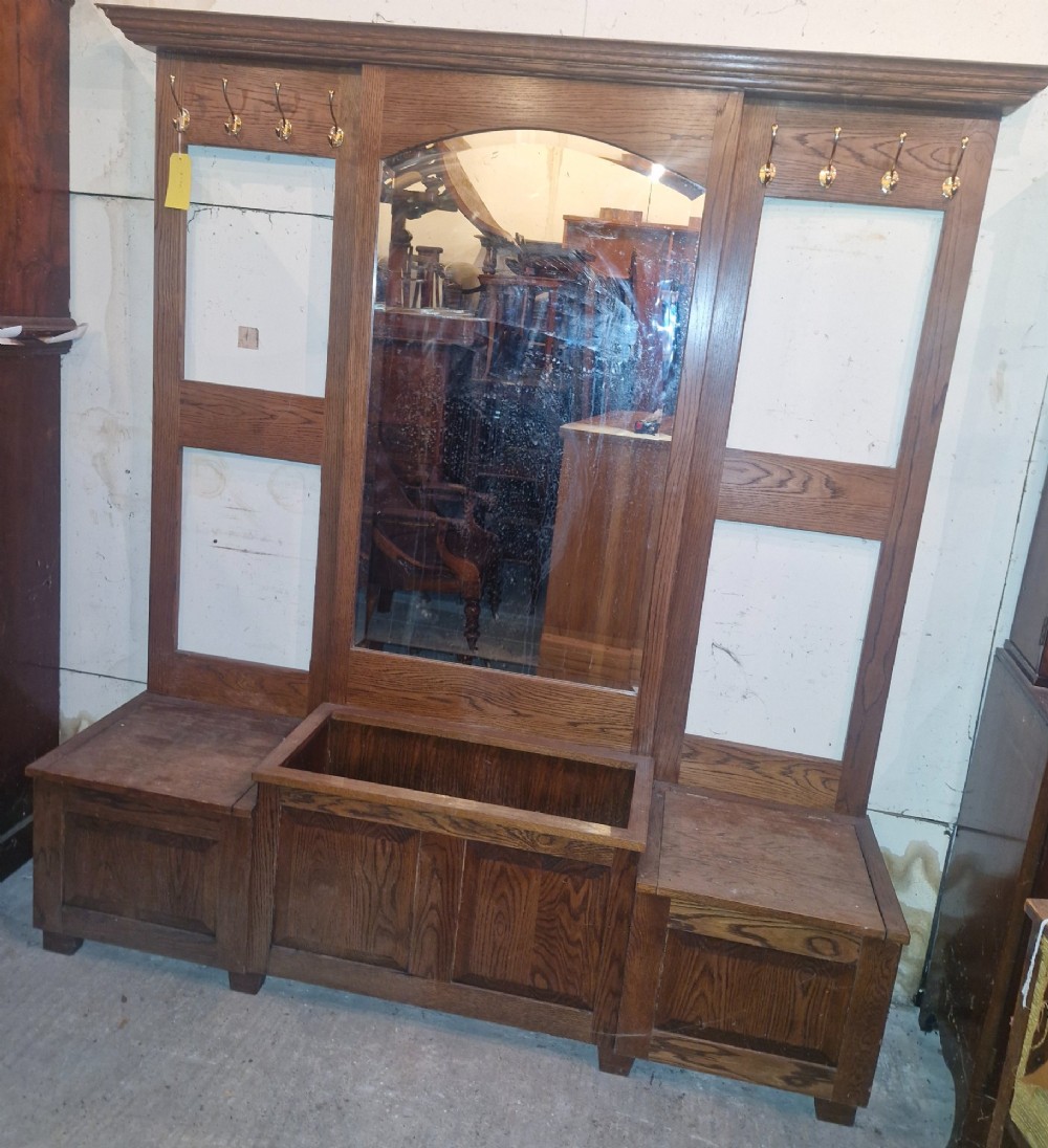 A large dark oak hall stand featuring a central arched mirror flanked by brass coat hooks, set above built-in storage boxes and a central trough base. View IX