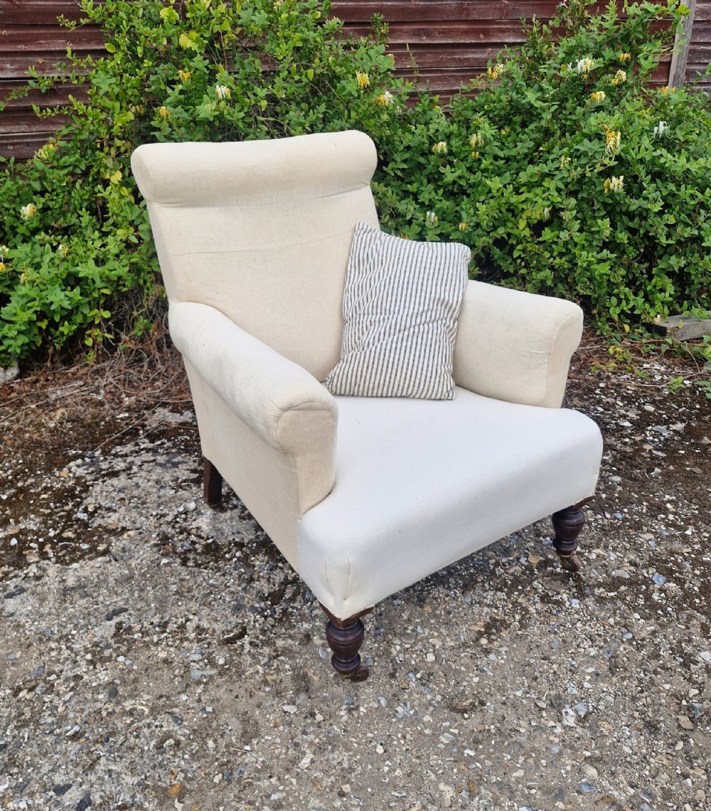 A late Victorian upholstered armchair in neutral cream fabric featuring a scrolled backrest, padded armrests, and turned mahogany front legs on brass castors. View II