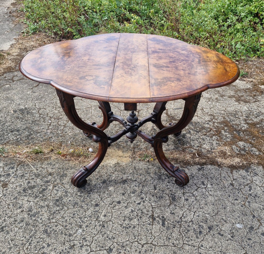A Victorian burr walnut scissor action table featuring a shaped oval top with distinct figure grain and carved cabriole legs with a central turned stretcher finial.