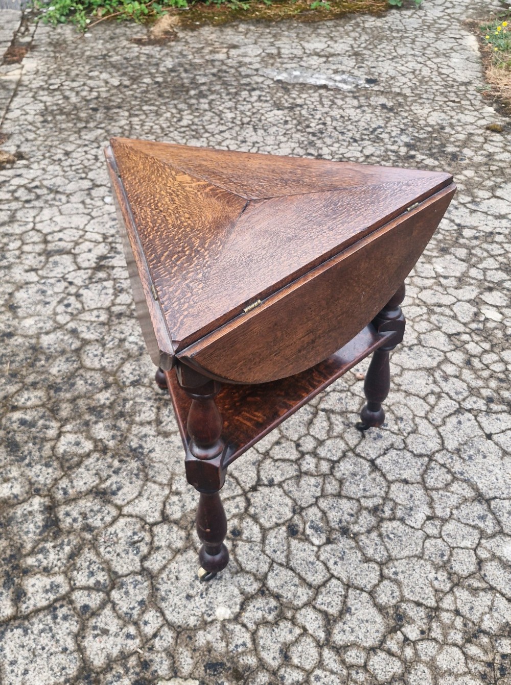 A dark oak three-legged cricket table featuring a drop-leaf circular top, a triangular lower shelf, turned legs, and brass casters. View II