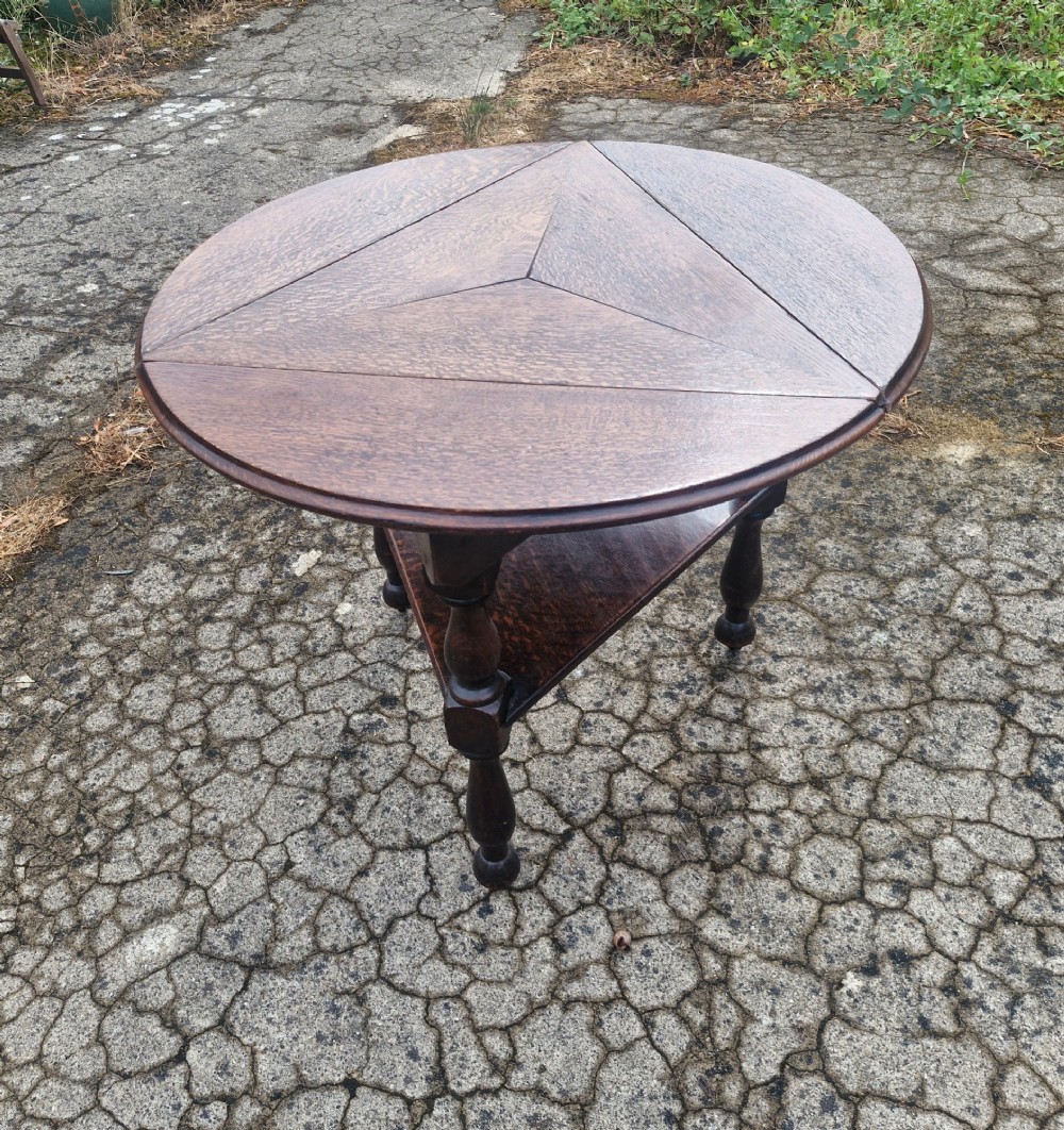 A dark oak three-legged cricket table featuring a drop-leaf circular top, a triangular lower shelf, turned legs, and brass casters. View III