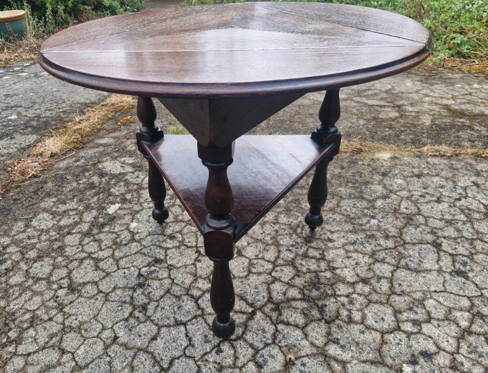 A dark oak three-legged cricket table featuring a drop-leaf circular top, a triangular lower shelf, turned legs, and brass casters. View IV
