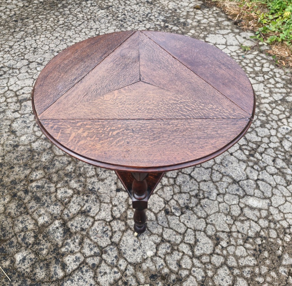 A dark oak three-legged cricket table featuring a drop-leaf circular top, a triangular lower shelf, turned legs, and brass casters. View V