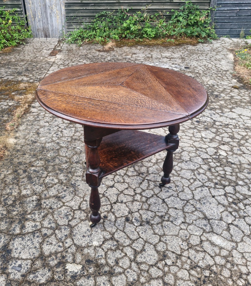 A dark oak three-legged cricket table featuring a drop-leaf circular top, a triangular lower shelf, turned legs, and brass casters. View VI