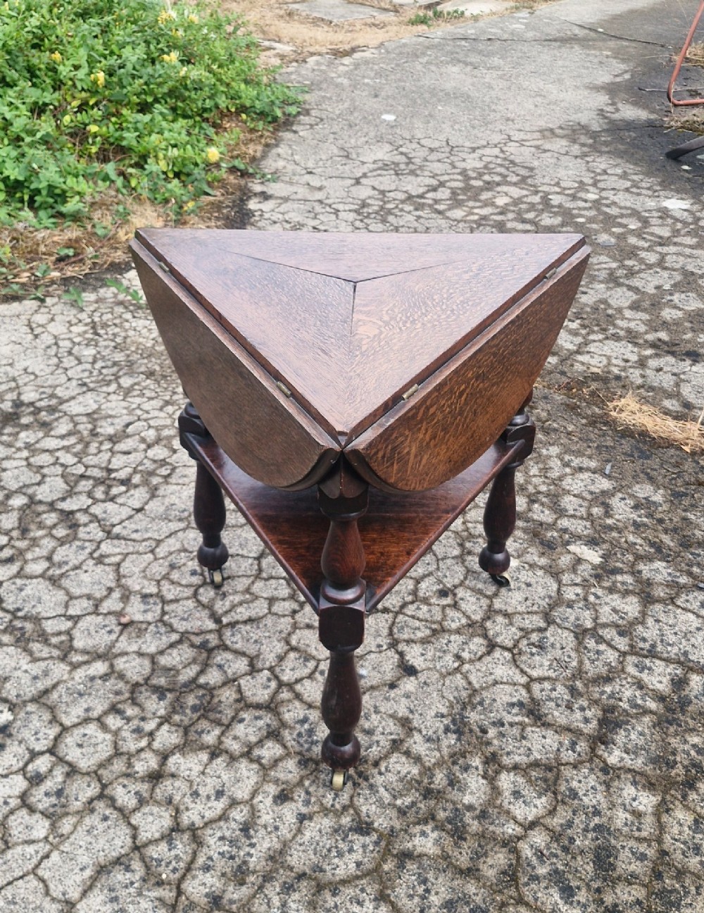 A dark oak three-legged cricket table featuring a drop-leaf circular top, a triangular lower shelf, turned legs, and brass casters. View VII