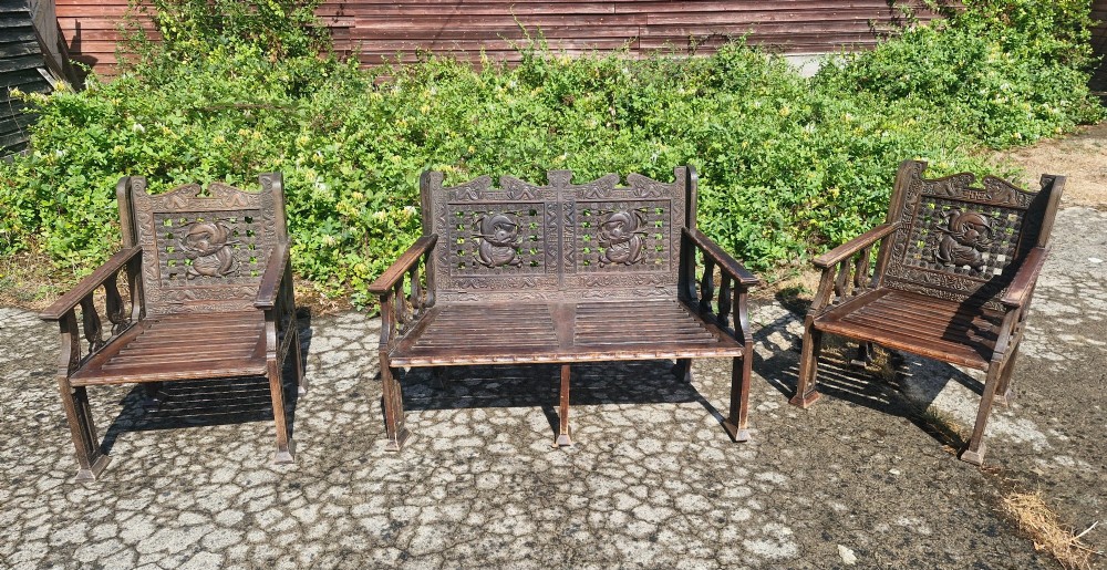 A three-piece dark carved teak seating suite comprising a double bench and two matching armchairs with intricate relief-carved backrests and slatted seats. View II