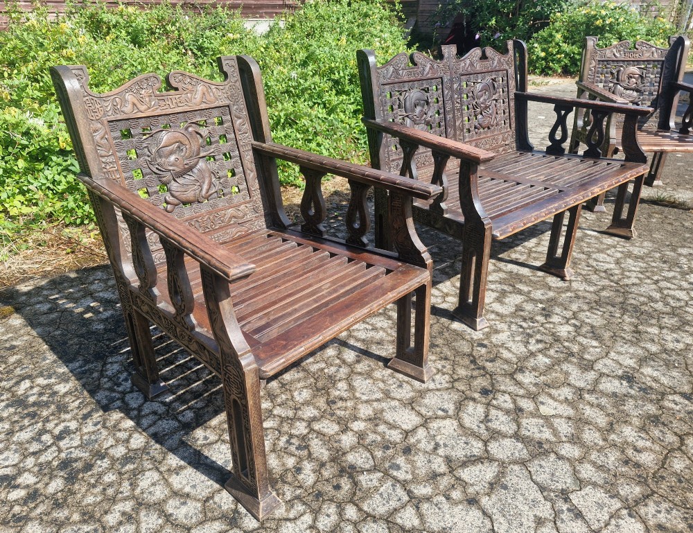 A three-piece dark carved teak seating suite comprising a double bench and two matching armchairs with intricate relief-carved backrests and slatted seats. View III