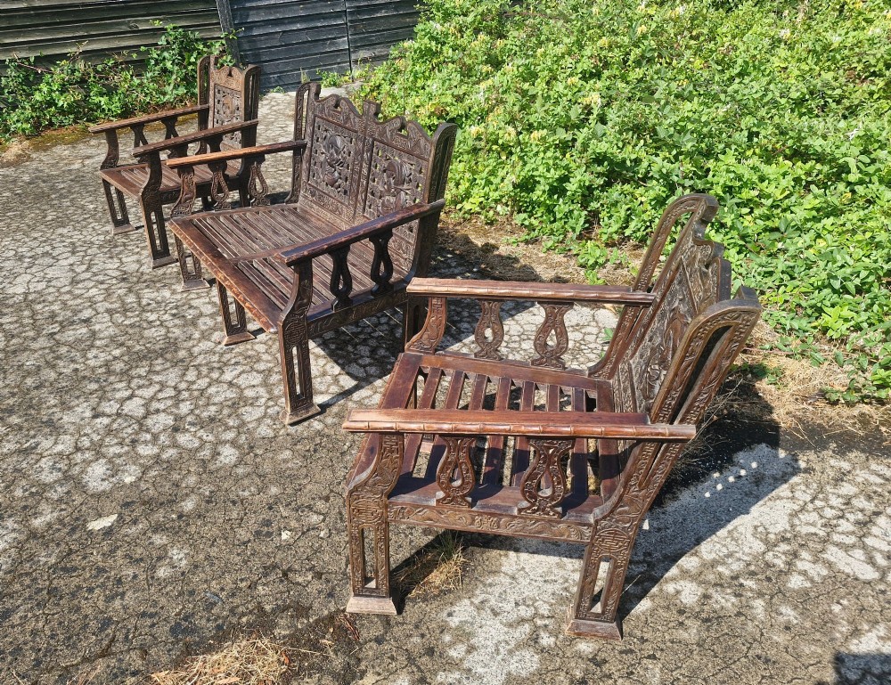A three-piece dark carved teak seating suite comprising a double bench and two matching armchairs with intricate relief-carved backrests and slatted seats. View V