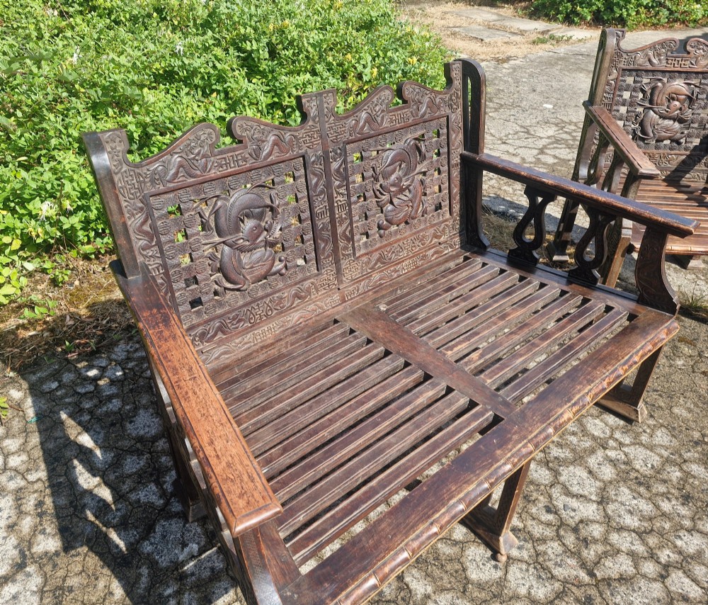A three-piece dark carved teak seating suite comprising a double bench and two matching armchairs with intricate relief-carved backrests and slatted seats. View VI