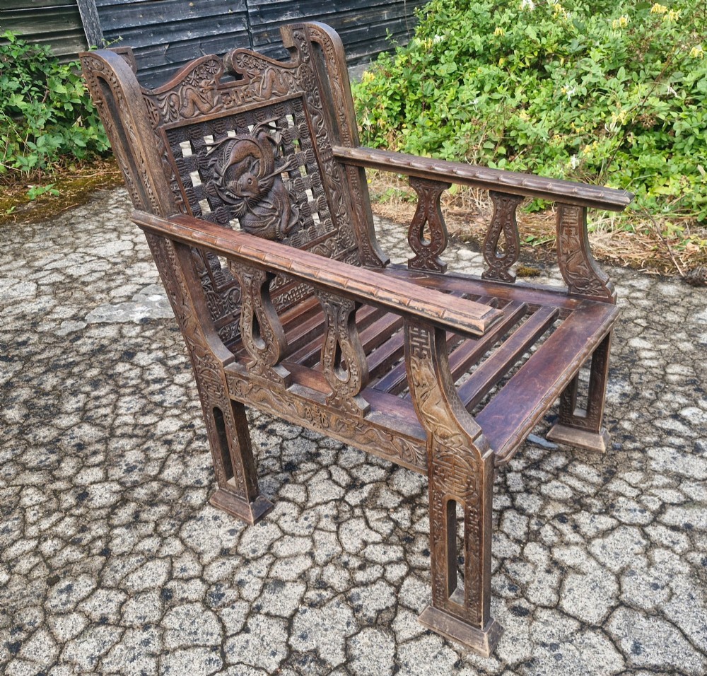 A three-piece dark carved teak seating suite comprising a double bench and two matching armchairs with intricate relief-carved backrests and slatted seats. View X