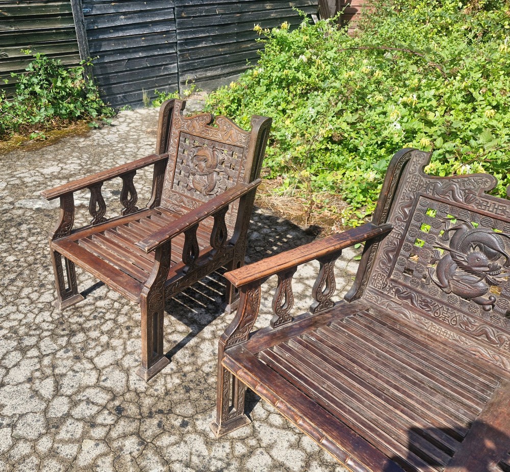 A three-piece dark carved teak seating suite comprising a double bench and two matching armchairs with intricate relief-carved backrests and slatted seats. View XI