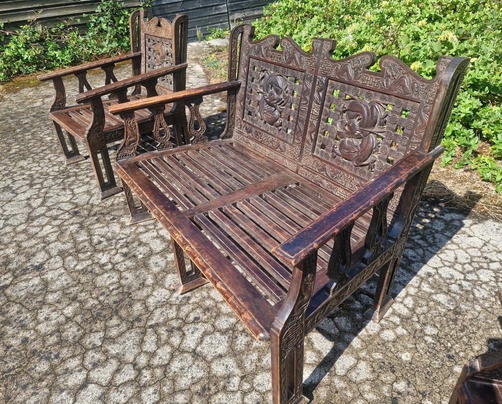 A three-piece dark carved teak seating suite comprising a double bench and two matching armchairs with intricate relief-carved backrests and slatted seats. View XII