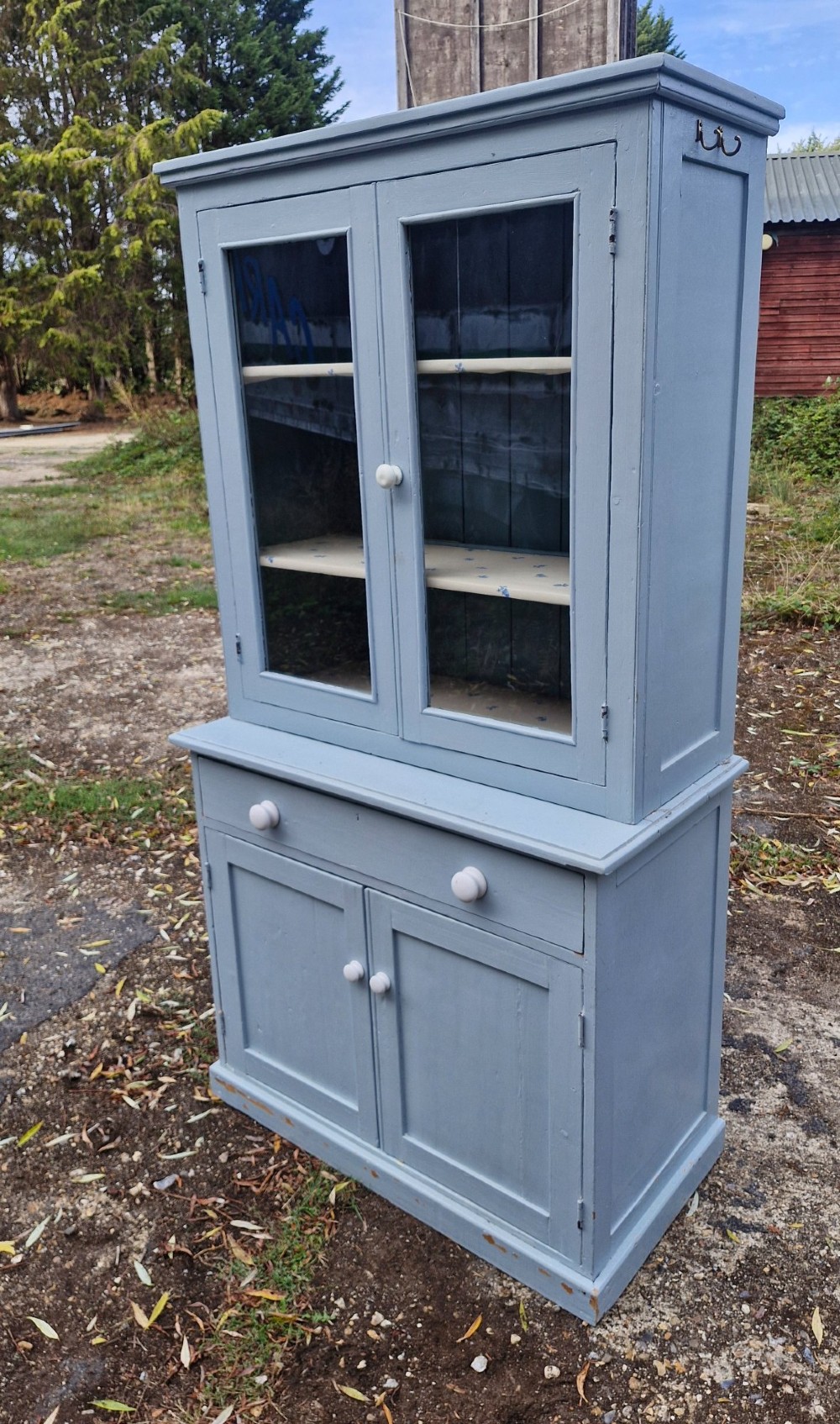 A freestanding two-part slate blue painted pine kitchen cupboard featuring an upper glazed display cabinet with two doors above a lower base with a drawer and panelled doors.