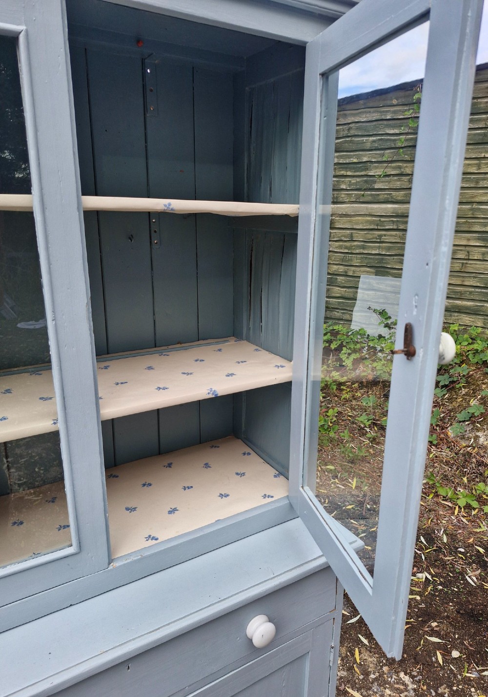 A freestanding two-part slate blue painted pine kitchen cupboard featuring an upper glazed display cabinet with two doors above a lower base with a drawer and panelled doors. View IV