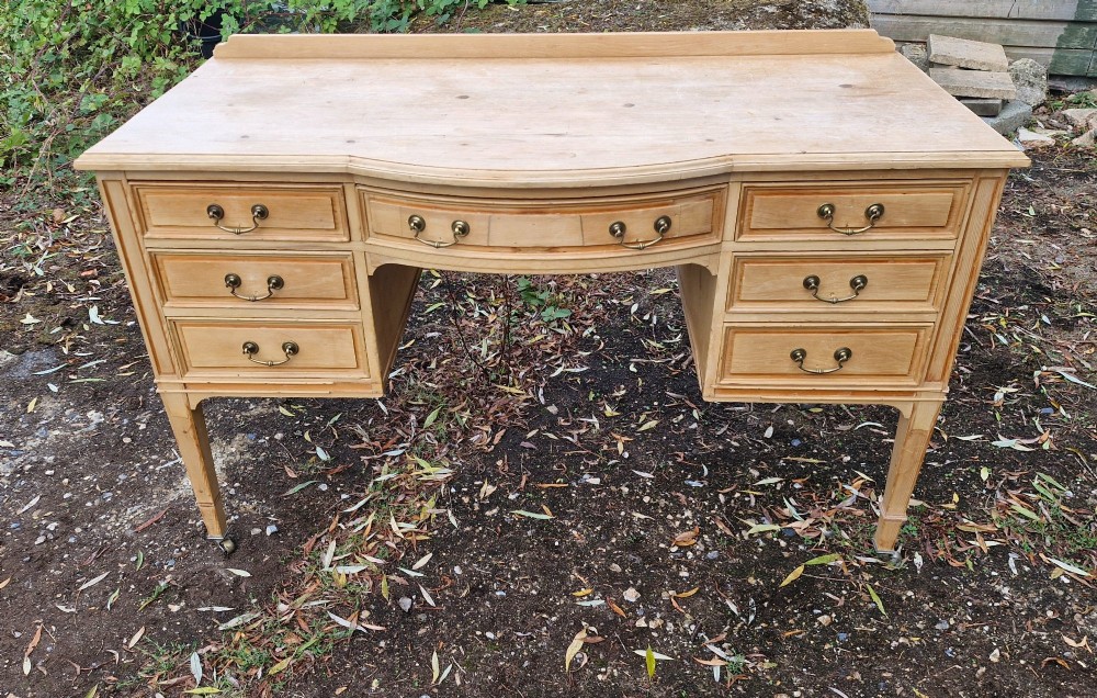 An Edwardian pine kneehole writing table featuring seven drawers with brass handles, a curved central drawer, and tapered legs on castors.
