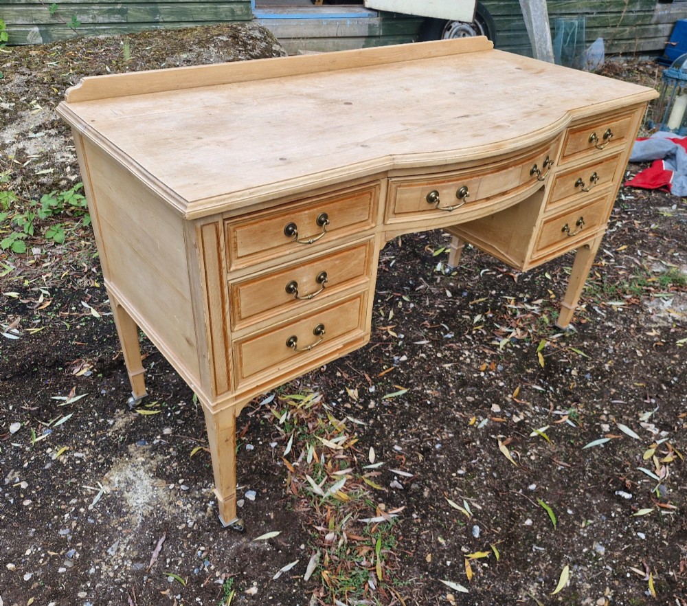 An Edwardian pine kneehole writing table featuring seven drawers with brass handles, a curved central drawer, and tapered legs on castors. View V