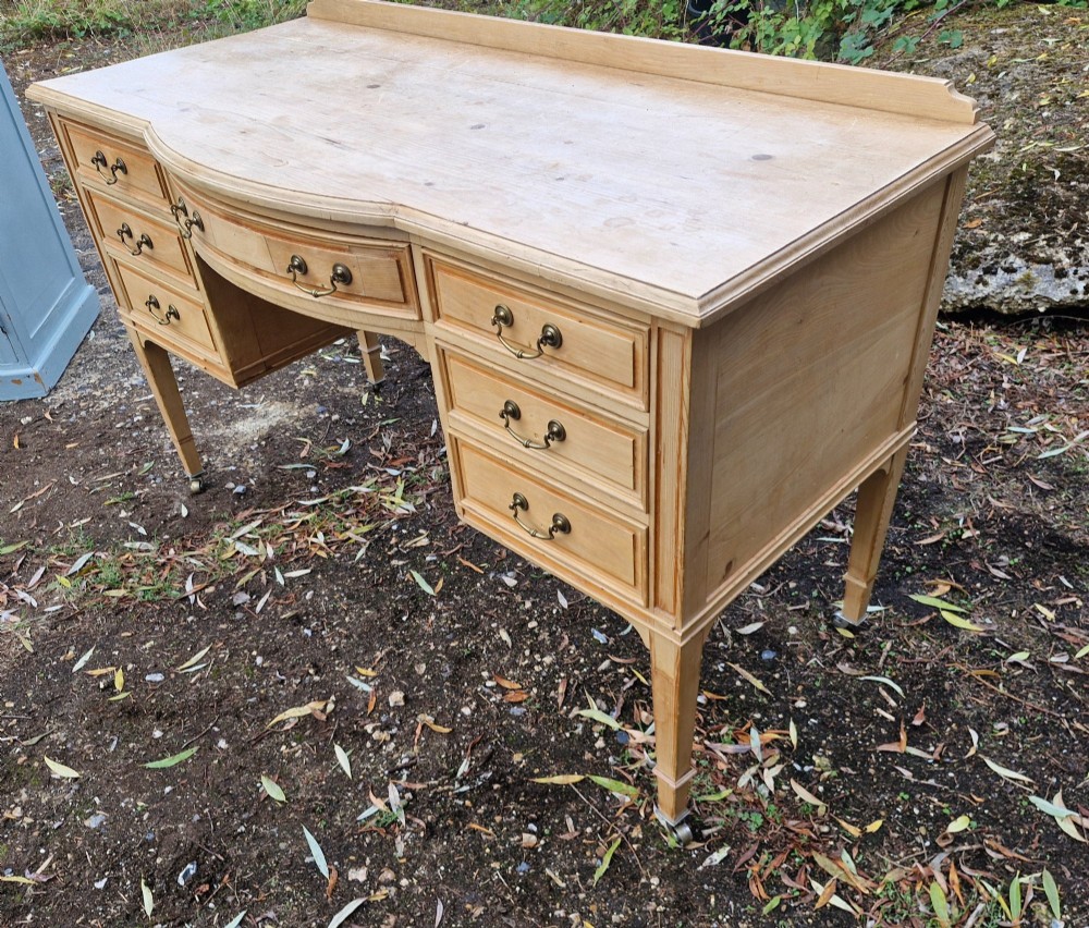 An Edwardian pine kneehole writing table featuring seven drawers with brass handles, a curved central drawer, and tapered legs on castors. View VI