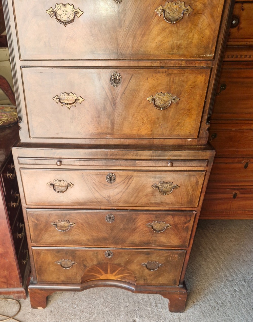 An early 20th century walnut chest on chest featuring brass drop handles, a pull-out brushing slide, and an inlaid sunburst motif on the lower drawer. View V