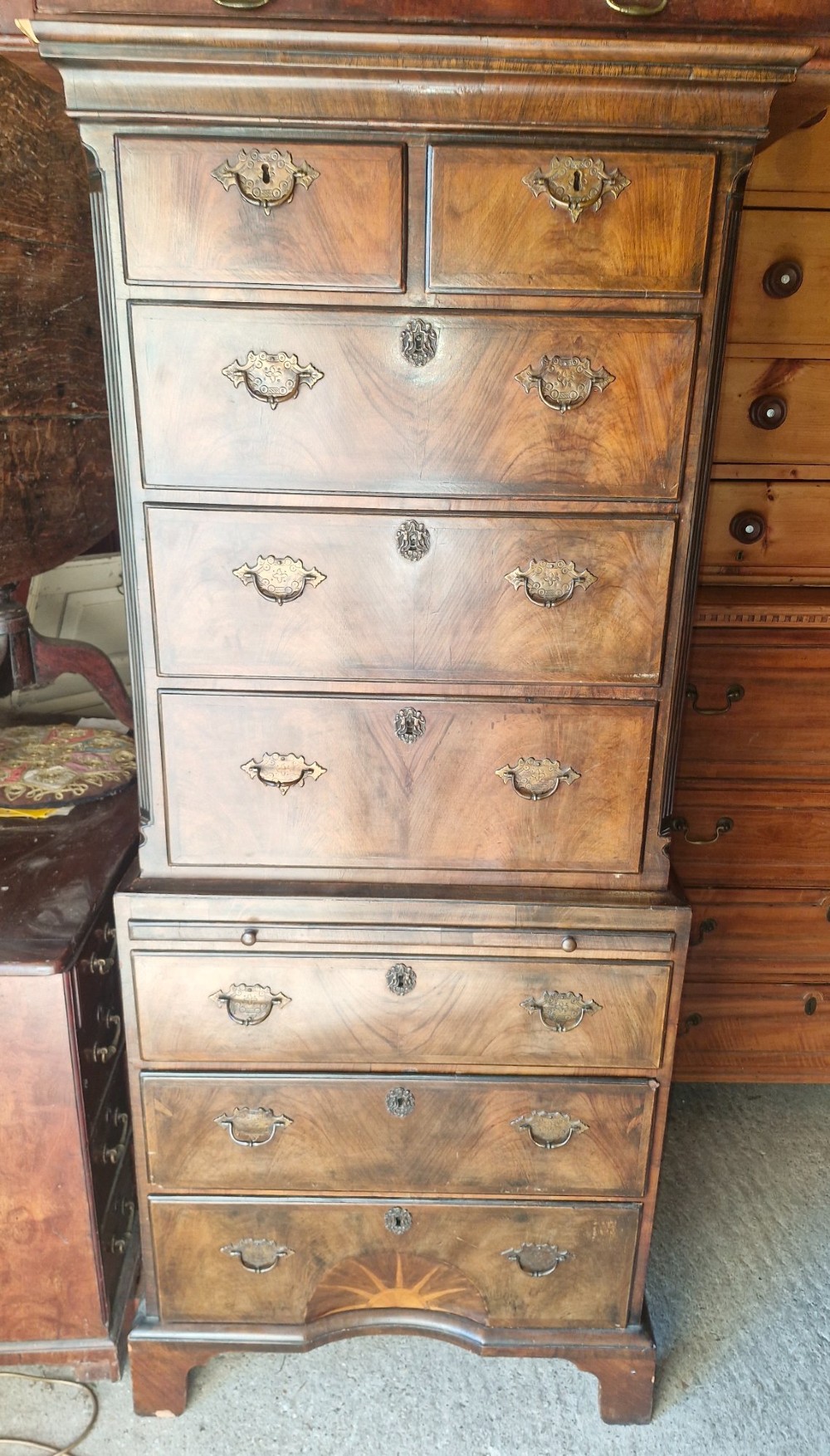 An early 20th century walnut chest on chest featuring brass drop handles, a pull-out brushing slide, and an inlaid sunburst motif on the lower drawer. View VI